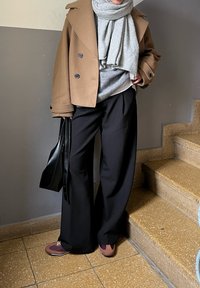 Brauner doppelreihiger Mantel über einem grauen Pullover, schwarze weit geschnittene Hose und kastanienbraune Sneaker. Schwarze Tasche in der Hand, stehend auf Treppen.