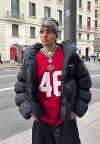 Jeune homme portant un bonnet camouflage, un maillot NFL rouge numéro 46, une grosse doudoune noire et un jean noir, debout dans une rue de la ville près d'un poteau.