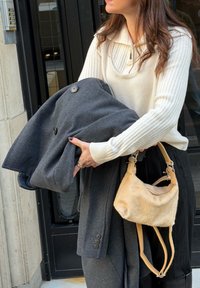 Γυναίκα που φοράει λευκό πουλόβερ κρατά διπλωμένο σκούρο γκρι παλτό και μια μπεζ τσάντα ενώ στέκεται κοντά σε μια μαύρη πόρτα.