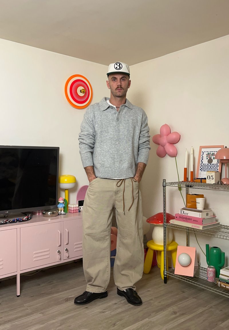 Maglione grigio chiaro a maglia con colletto, pantaloni beige a gamba larga e scarpe nere. La stanza presenta un arredamento colorato, una TV e un'unità di stoccaggio rosa.