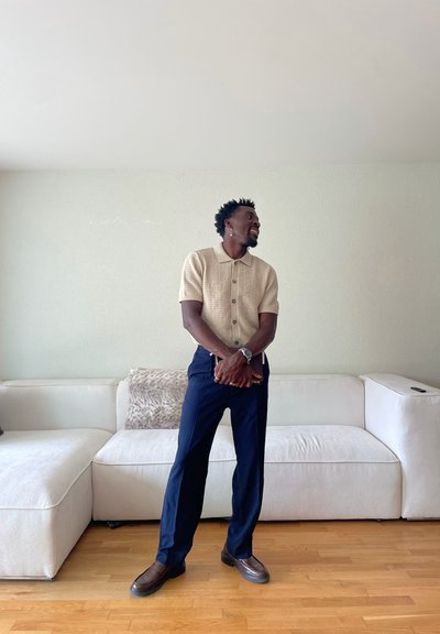 Hombre con camisa de punto beige de manga corta y pantalones azul marino, sosteniendo un libro, de pie y sonriendo frente a un sofá seccional blanco sobre un suelo de madera.