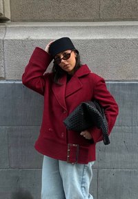 Manteau en laine rouge à double boutonnage avec poignets côtelés et détail boucle argentée, associé à un sac à main tressé noir et un chapeau noir. Jean en denim bleu clair.