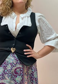 Schwarzer Weste mit Knöpfen, kombiniert mit einem weißen, gerafften Hemd. Der Rock hat ein buntes Paisleymuster und ist mit einer herzförmigen Gürtelschnalle verziert.
