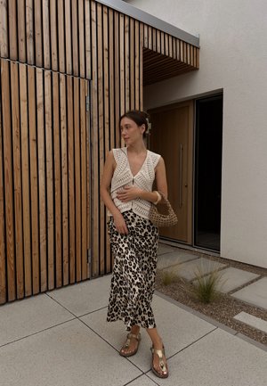Chaleco de ganchillo en marfil, falda maxi de satén con estampado de leopardo y sandalias doradas. Un bolso de paja añade textura. Fondo de madera moderna.