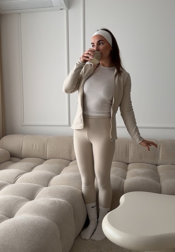 Vrouw in beige sportkleding en witte hoofdband die uit een mok drinkt, staand naast een zachte bank en een lichtgekleurde tafel in een minimalistische kamer.