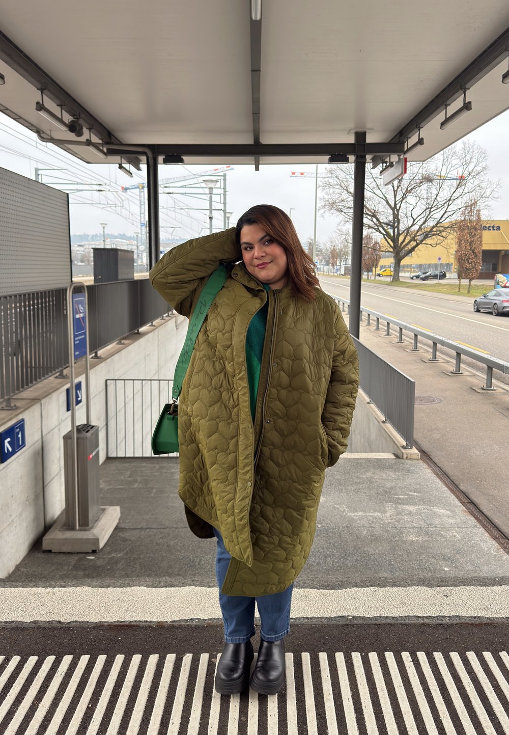 Vihreä vuorattu takki suurilla timanttimaisilla kuvioilla, päällä vihreä toppi, yhdistetty sinisiin farkkuihin ja suurikokoisiin mustiin saappaisiin. Mukana vihreä laukku.