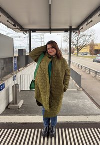 Vihreä vuorattu takki suurilla timanttimaisilla kuvioilla, päällä vihreä toppi, yhdistetty sinisiin farkkuihin ja suurikokoisiin mustiin saappaisiin. Mukana vihreä laukku.