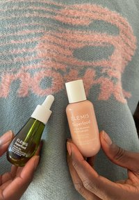 Hände halten zwei Elemis Superfood Hautpflegeprodukte: ein grünes Gesichtsöl mit Pipette und eine Flasche mit pfirsichfarbenem Glow-Priming-Moisturizer.