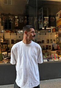 O persoană îmbrăcată cu un tricou alb larg stă în fața unei vitrine de magazin care afișează diferite discuri muzicale și mărfuri.