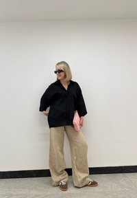 Chemise en lin noire à boutons; pantalons beige à jambes larges; pochette tissée rose; baskets brunes et blanches; lunettes de soleil noires rondes.