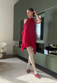 Rotes, rüschendes Kleid mit hohem Kragen, transparenter Stoff und schichtiger Gestaltung. Kombiniert mit glänzenden roten spitzen Ballerinas. Inneneinstellung mit grünen Akzenten.