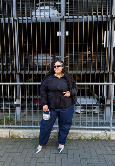 Chaqueta negra con cintura ajustable, combinada con jeans azules. Tacones plateados de punta y un pequeño bolso metálico completan el look, todo en un aparcamiento.