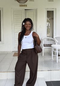 Femme souriante aux longs cheveux bouclés et fleur blanche, portant un débardeur blanc et un pantalon marron, tenant un sac tissé marron sur le porche avec des chaises blanches.