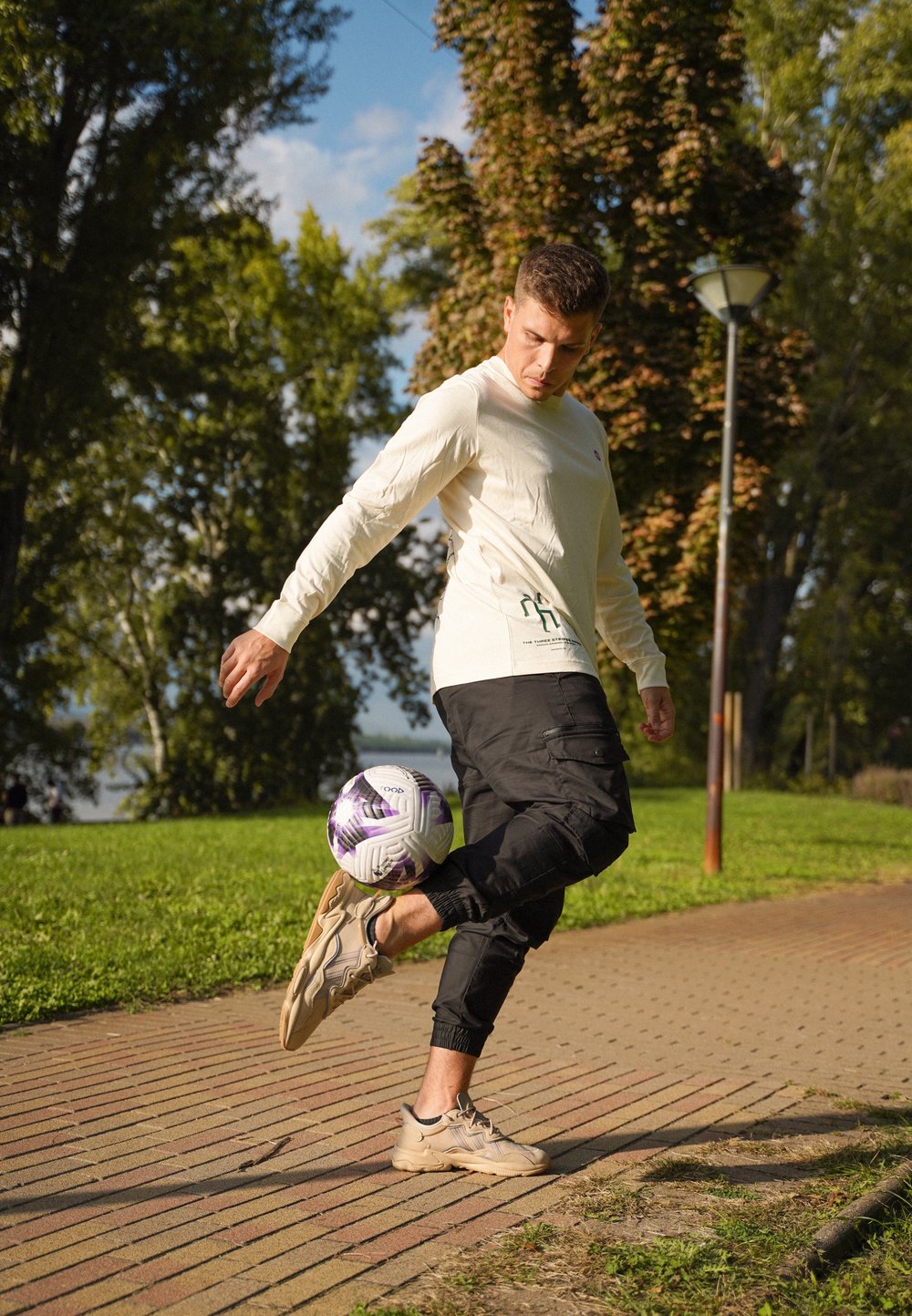 Ανοιχτό μπεζ πουκάμισο μακρύ μανίκι με πράσινη εκτύπωση και μαύρο cargo παντελόνι; κάνει ζογκλερικά με μια λευκή και μωβ μπάλα ποδοσφαίρου σε ένα πλακόστρωτο μονοπάτι σε ένα πάρκο.