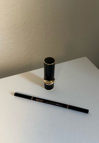 Svart läppstiftstub med gyllene detaljer bredvid en svart ögonbrynspenna märkt "BROW WIZ" och "ANASTASIA." Släta texturer, minimalistisk design.