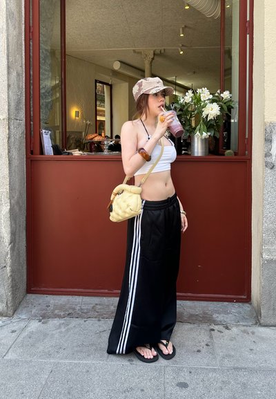 Pantalones de chándal negros con rayas blancas, una camiseta blanca corta, una gorra beige con un patrón y un bolso amarillo. Sosteniendo una bebida cerca de flores.