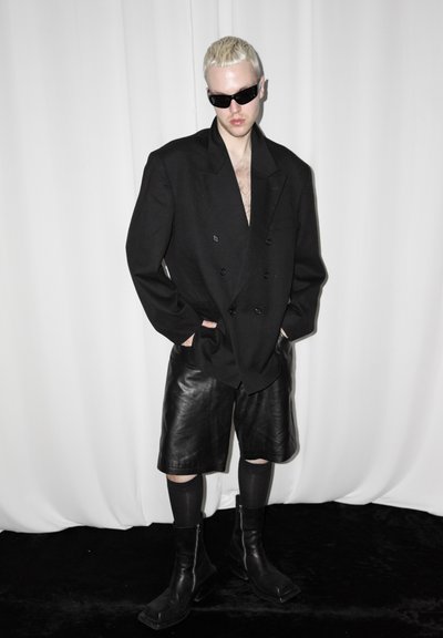 Persona con cabello rubio corto y gafas de sol lleva una blazer negra de gran tamaño, pantalones cortos de cuero, calcetines negros y botas negras contra fondo blanco.