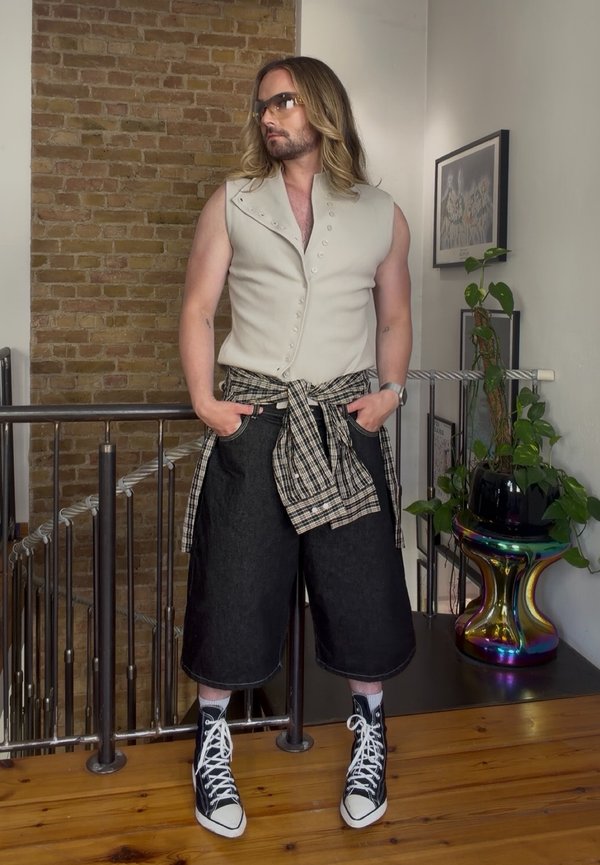 Persoon met lang haar, draagt een mouwloos beige vest, geruit hemd om de taille geknoopt, brede zwarte shorts en hoge sneakers, staand binnen.