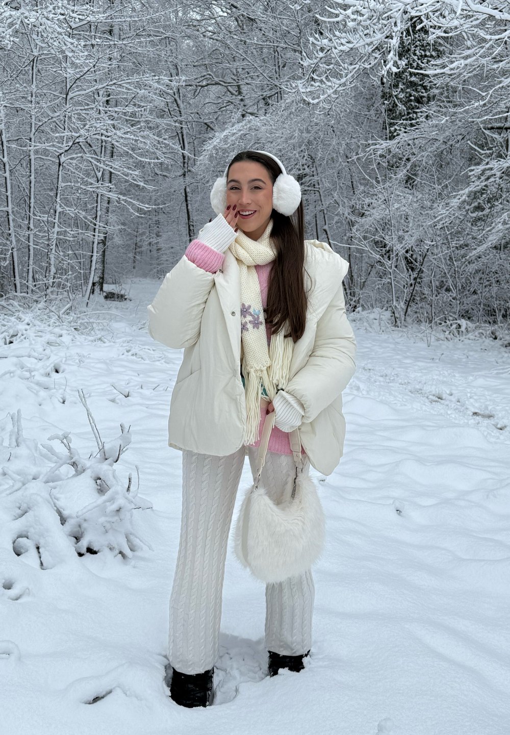 Jaqueta puffer branca, suéter rosa, calças brancas de malha, cachecol creme, earmuffs peludos e uma bolsa felpuda branca numa paisagem nevada.