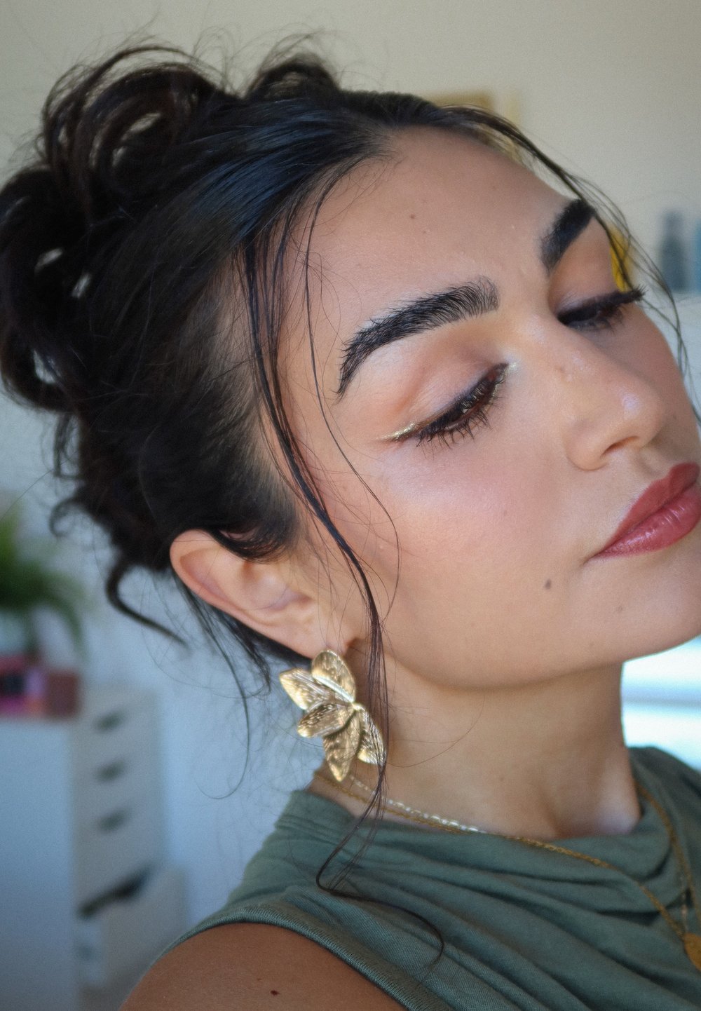 Orecchini a forma di foglia d'oro, top senza maniche di colore verde oliva, trucco sottile con un leggero splendore, capelli scuri acconciati in ricci morbidi.