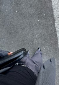 Sac à main noir tenu par une personne portant un haut noir ajusté et un pantalon gris taillé, surmonté d'un blazer gris à rayures, se tenant sur le pavé.