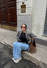 Indossa un blazer oversize a fantasia, una maglietta nera e jeans azzurri chiari, seduta su gradini di pietra con una borsa marrone borchiata e sneakers grigie.