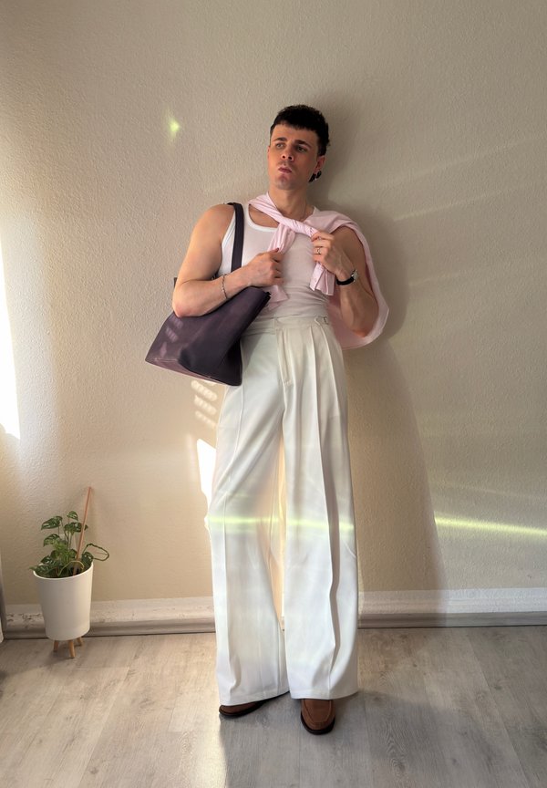 Uomo che indossa pantaloni bianchi a gamba larga, top bianco senza maniche, maglione rosa sulle spalle, tiene una borsa nera, in piedi all’interno vicino a un muro beige.
