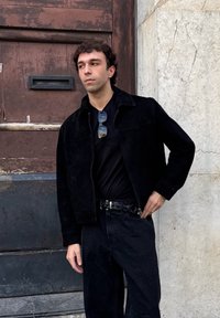 Jachetă din suede negru cu un design cu guler, purtată cu o cămașă neagră și blugi. Persoana poartă ochelari de soare și pozează în fața unei pereți texturat.