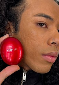 Contenitore cosmetico ovale rosso etichettato "TIRTIR" tenuto contro una superficie della pelle texture; la persona ha capelli ricci neri e indossa una giacca nera con zip.