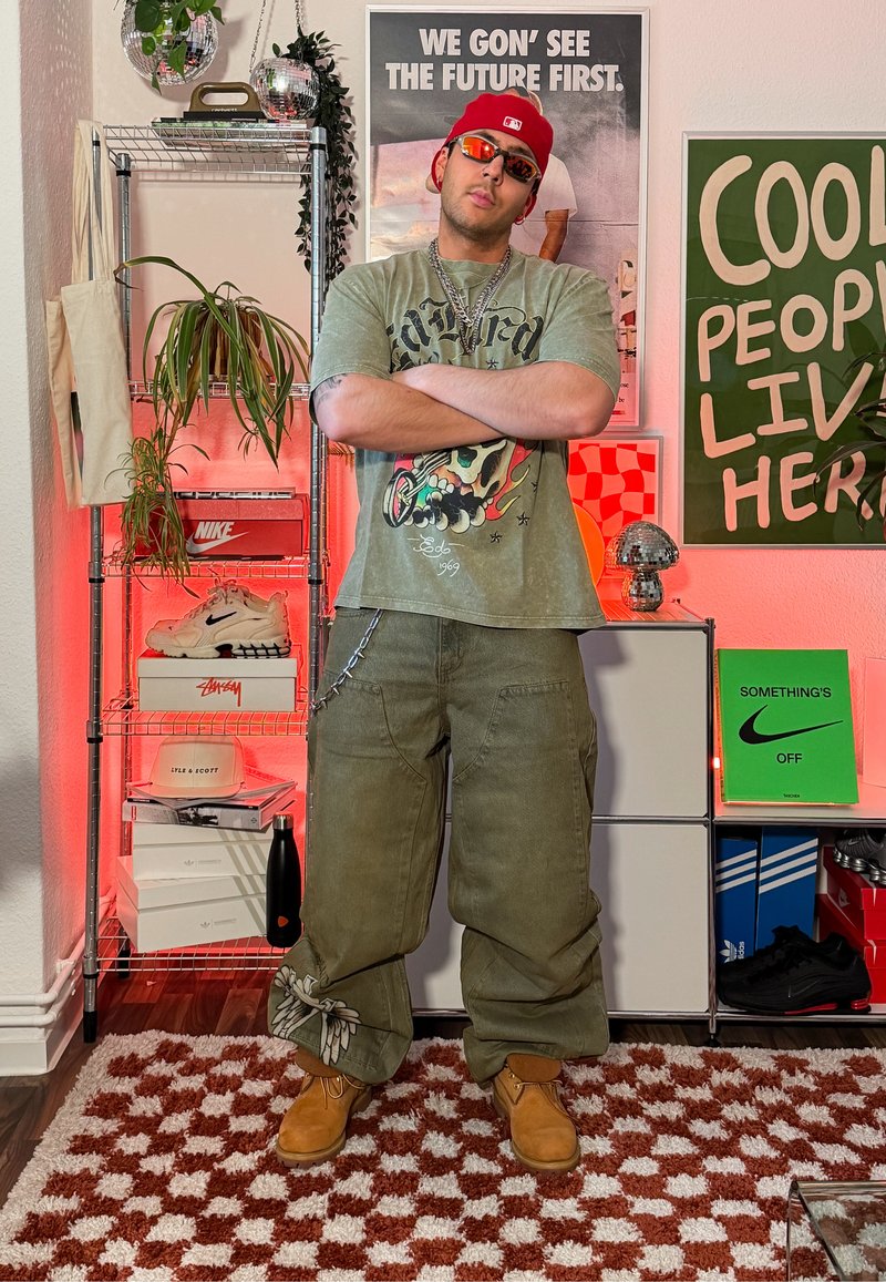 Jeune homme en tee-shirt graphique vert et pantalon cargo, les bras croisés, dans une pièce avec des affiches, des plantes, des boîtes à chaussures et un éclairage rouge.
