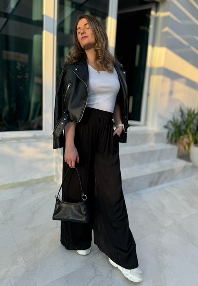 Chaqueta de motorista de cuero negra, camiseta blanca ajustada, pantalones de pierna ancha negros, bolso pequeño negro y zapatillas blancas sobre una superficie de mármol.
