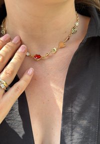 Χρυσό κολιέ σε σχήμα καρδιάς με κόκκινες και διάφανες πολύτιμες πέτρες, εναλλασσόμενες με χρυσά στοιχεία. Φορεμένο κοντά στον λαιμό, αναδεικνύει το σχέδιο.