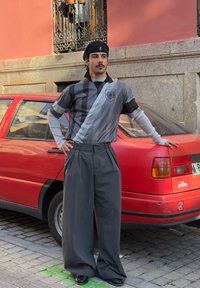 Top scurt cu dungi gri și negre, cu logo, pantaloni largi gri și o beretă neagră, stând lângă o mașină roșie.