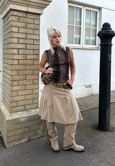 Top sin mangas de estampado de leopardo, falda acampanada beige con flecos, pantalones y botas beige a juego. Los accesorios incluyen un bolso pequeño marrón y gafas de sol.