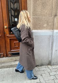 Brown wool-blend coat with belt, paired with light blue flared jeans and black leather boots. Black textured bag slung over shoulder.