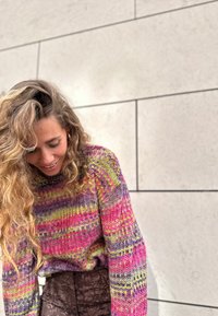 Mikina z příze v mnoha barvách s živými pruhy v růžové, žluté a fialové barvě, s volným střihem a žebrovaným výstřihem, spárovaná s texturovanými hnědými kalhotami.