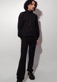 Personne avec des cheveux bouclés portant un blouse noir transparent à col haut, un pantalon noir et des chaussures à plateforme noires se tenant devant un fond uni.
