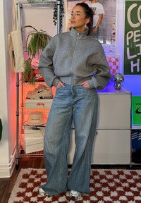 Jeune femme vêtue d'un sweat-shirt gris à fermeture éclair et d'un jean à jambes larges, debout à l'intérieur, les mains dans les poches, à côté d'étagères contenant des baskets et des plantes.