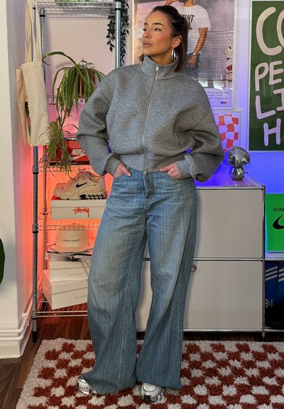 Joven mujer con sudadera gris con cremallera y vaqueros de pierna ancha, de pie en el interior con las manos en los bolsillos, junto a estanterías con zapatillas y plantas.