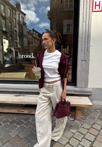 Bordeaux rits-hoodie, witte aansluitende top, brede beige geruite broek en een kastanjebruine handtas van gevlochten materiaal. Houdt een doorzichtige beker met een rietje vast.