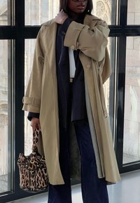 Beige oversized trenchcoat med vida ärmar, knappar och en slät textur, kombinerad med en leopardmönstrad väska med gyllene detaljer.