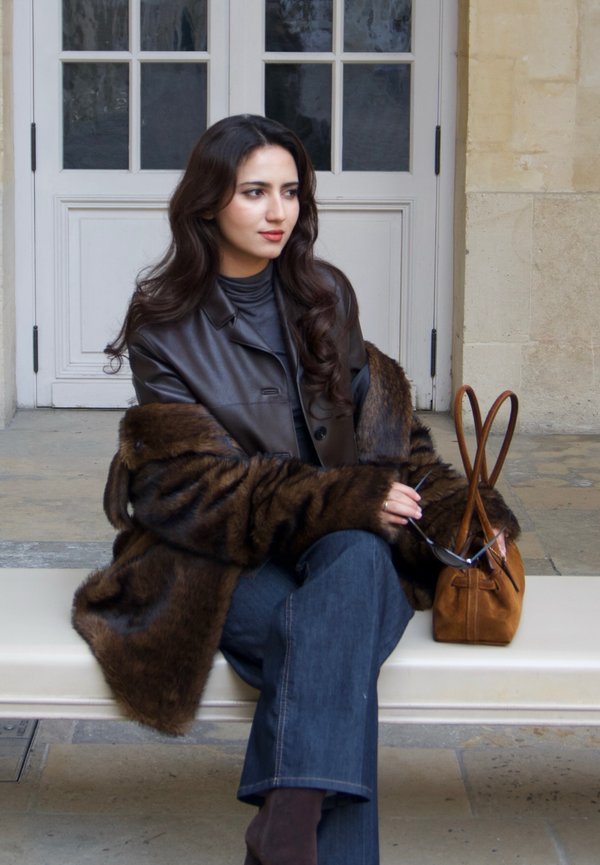 Braune Lederjacke über einem dunklen Rollkragenpullover, kombiniert mit hellblauen Jeans. Brauner Kunstpelzmantel, braune Handtasche und Sonnenbrille in der Hand.