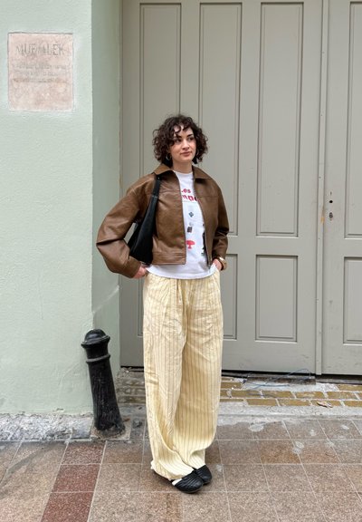 Chaqueta de cuero marrón recortada, camiseta gráfica blanca, pantalones de pierna ancha a rayas color crema y zapatos negros tipo slip-on, de pie frente a una pared verde clara.