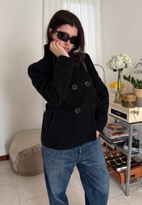 Γυναίκα με μαύρο παλτό και γυαλιά ηλίου ποζάρει εσωτερικά κοντά σε ράφι με φυτά, βιβλία και τραπέζι ποδοσφαίρου.