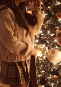 Sweater de malha bege com um corte descontraído e bainha canelada, combinado com uma saia xadrez em tons de castanho. O fundo apresenta uma árvore de Natal decorada.