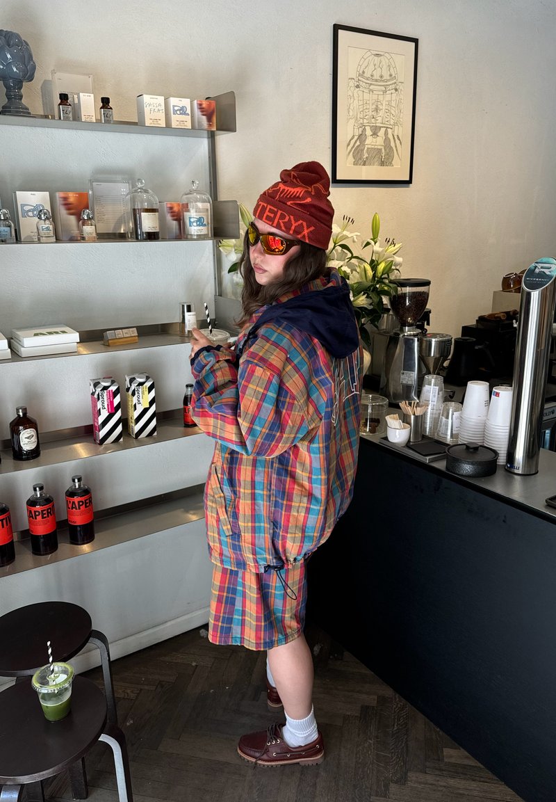 Personne en veste à carreaux colorés et short, bonnet rouge et lunettes de soleil tenant une tasse dans un café moderne près des étagères avec des boissons.