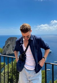 Casual outfit featuring a navy blue button-up shirt, a white tank top, light blue shorts, and sunglasses. Scenic ocean backdrop with boats.