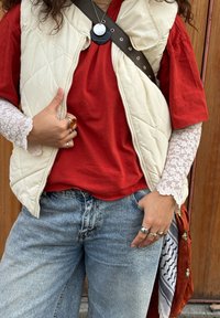 Bēša izšūta veste pār sarkanu T-kreklu, kārtainas mežģīņu garās piedurknes, gaiši zilas džinsas un vairāki gredzeni; brūna plecu siksna ar melnu piekariņu.