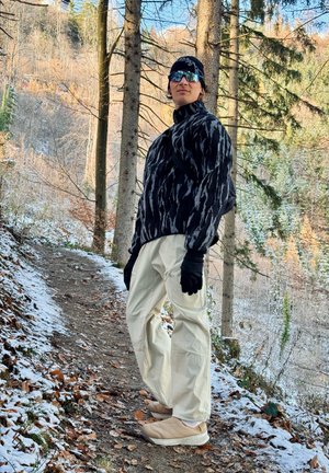 Una persona está de pie en un sendero nevado, vistiendo una chaqueta de forro polar negra y gris con estampado, pantalones crema, guantes negros y zapatos claros.