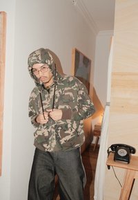 Sweat à capuche à motif camouflage avec cordons et accents bruns, associé à un jean denim foncé. La pièce présente des éléments en bois et un téléphone rétro.